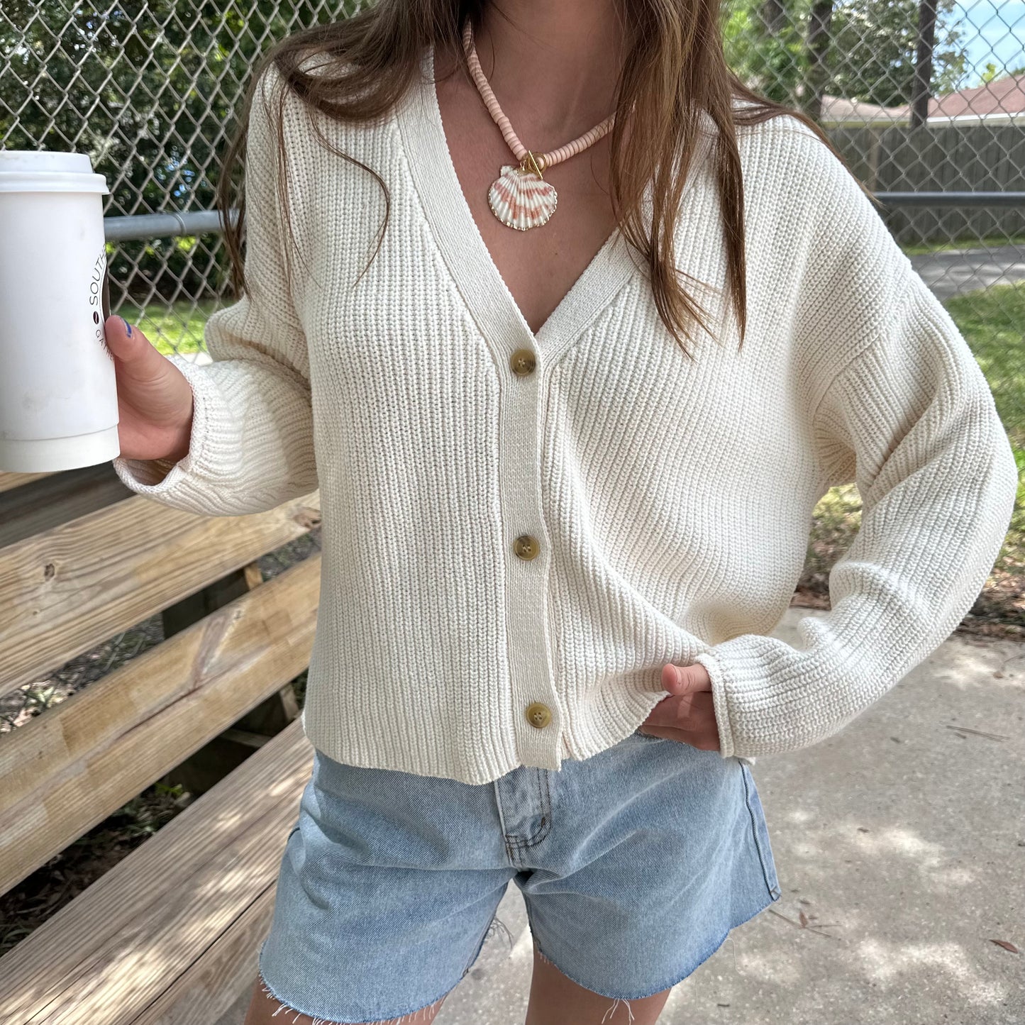 Woman wearing chunky cocoon cardigan, Ivory knit cardigan sweater and seashell necklace holding a coffee.