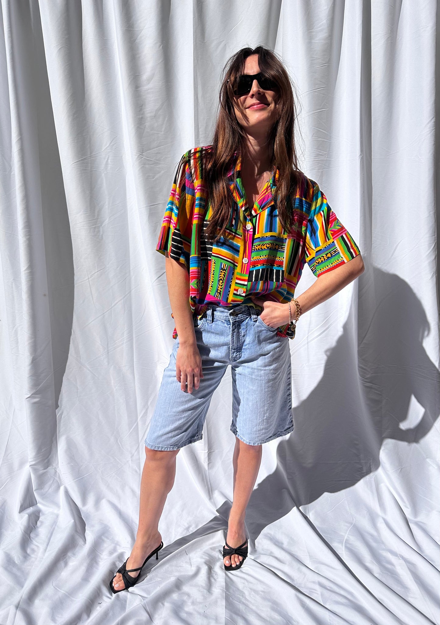 Girl standing in front of white backdrop wearing colorful printed vintage blouse and long light wash vintage denim shorts. 