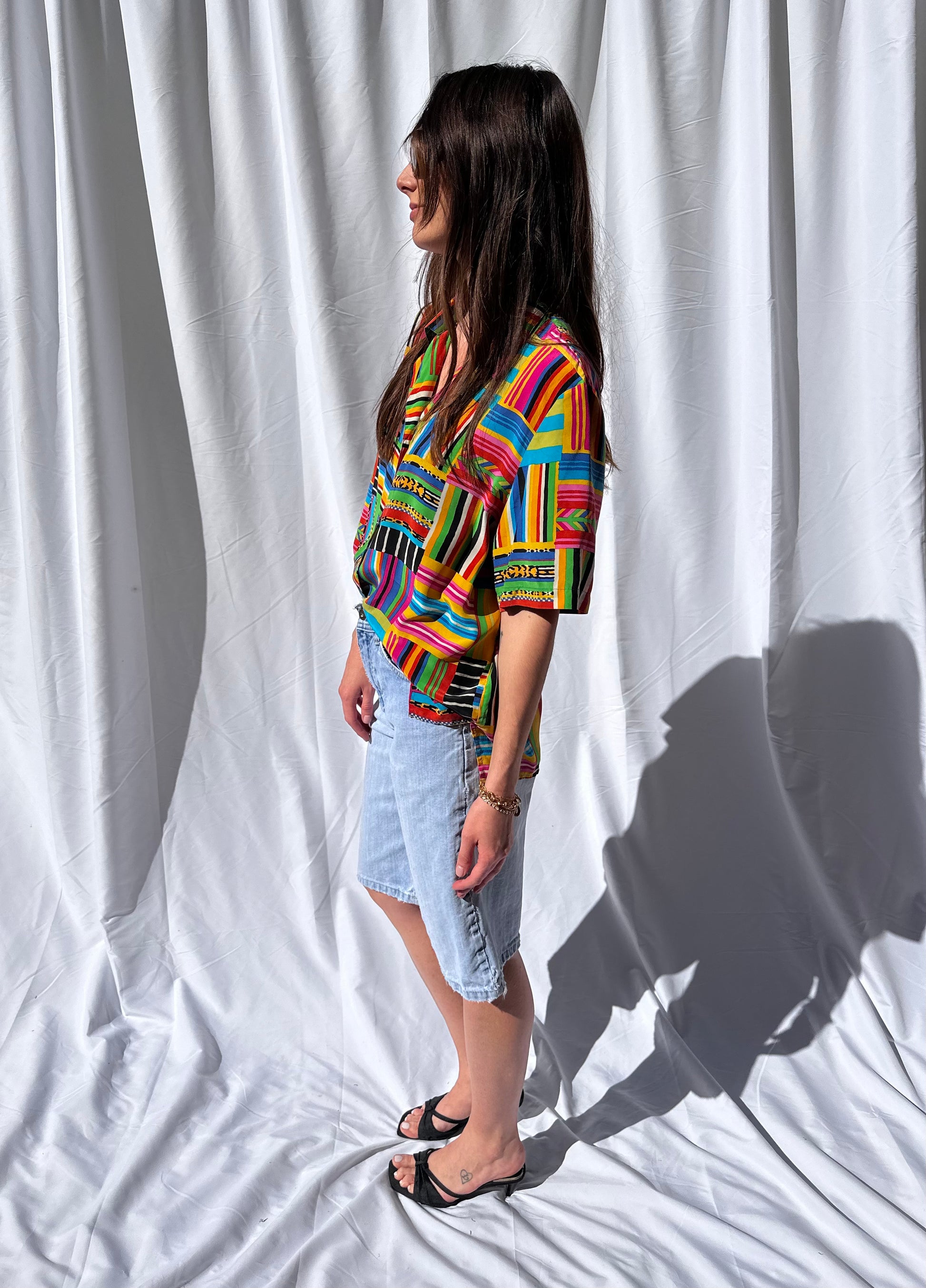 Girl standing in front of white backdrop wearing colorful printed vintage blouse and long light wash vintage denim shorts. 