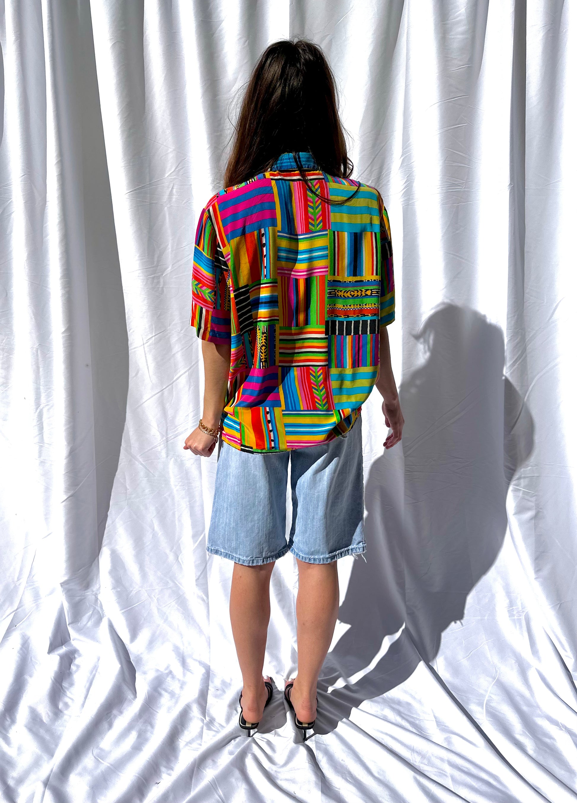 Girl standing in front of white backdrop wearing colorful printed vintage blouse and long light wash vintage denim shorts. 