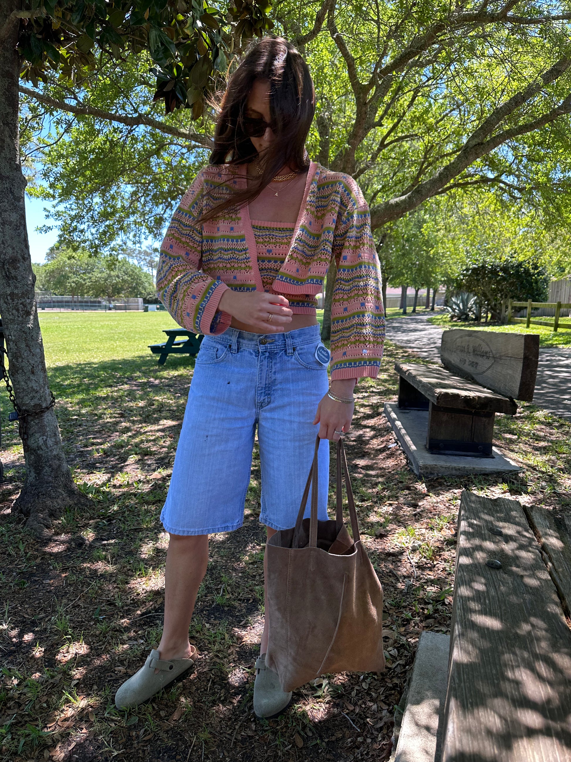 Girl in park wearing long vintage denim shorts holding tan suede tote bag. Wearing crochet colorful cardigan and top. 