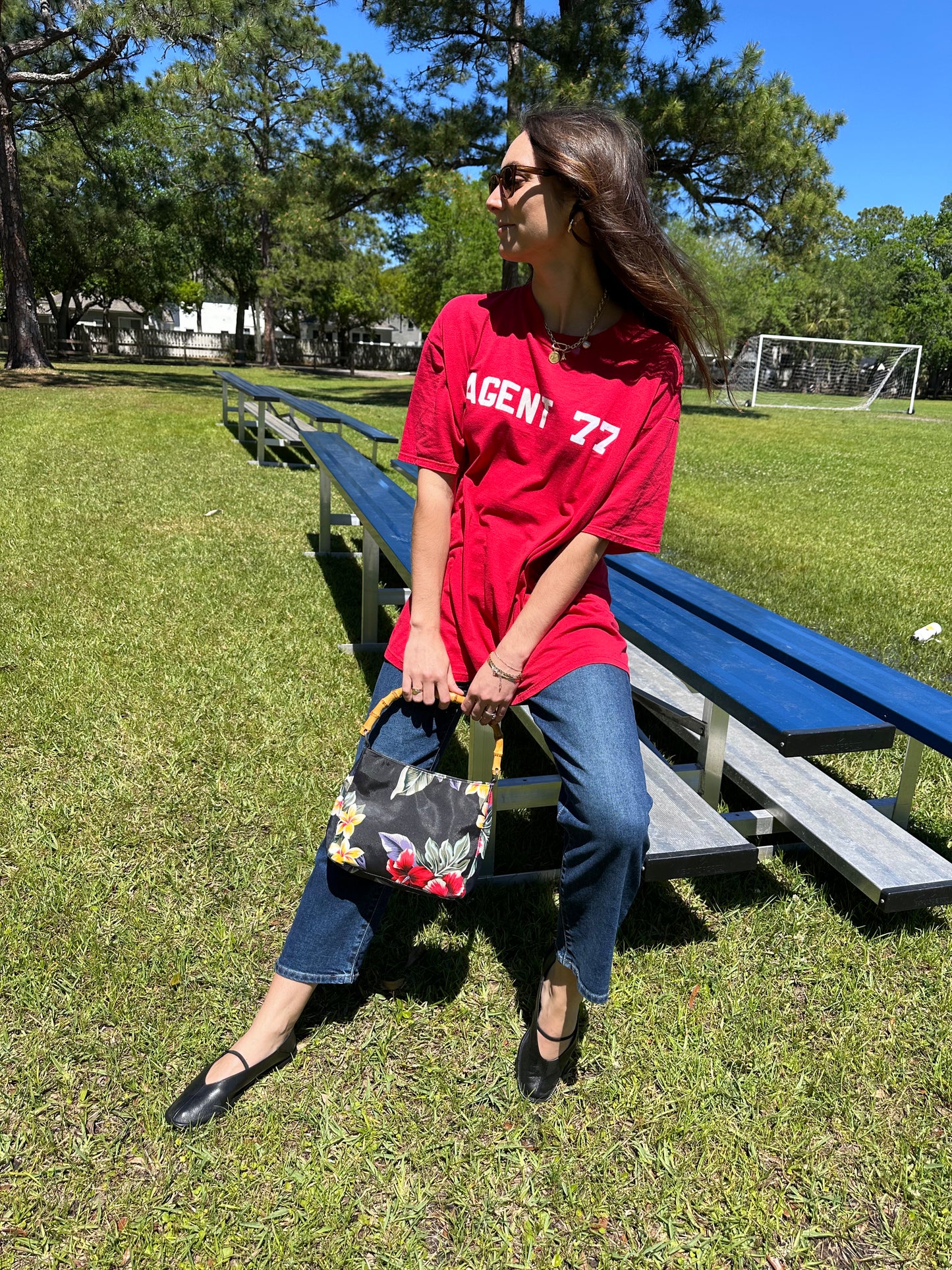 Girl sitting on bleachers wearing vintage red graphic tee and levis ribcage straight in mid wash holding vintage hawaiian print purse with bamboo handle. 